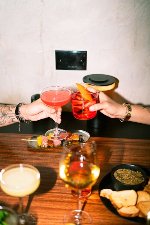 Two people clinking cocktail glasses over appetizers